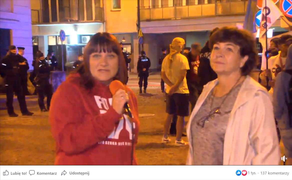Zrzut ekranu z Facebook Watch, w którym zamieszczono nagranie z Justyną Sochą i dr Dorotą Sienkiewicz. Na nagraniu widać obie kobiety przeprowadzające rozmowę wieczorną porą na tle grupy osób z transparentami oraz policji.
