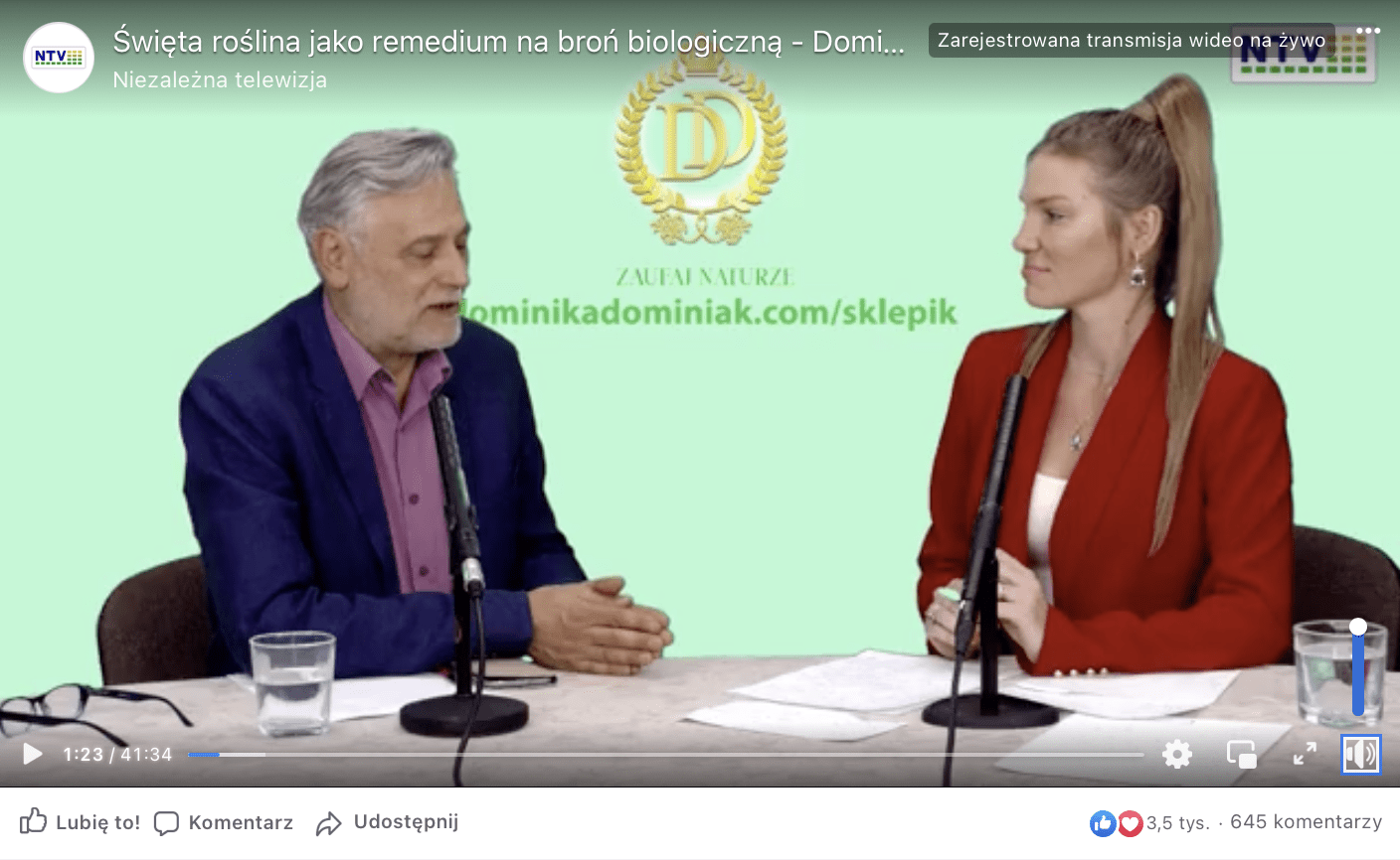 Zrzut ekranu kadru filmu udostępnionego na Facebooku. Na grafice widzimy mężczyznę ubranego w garniturę i koszulę oraz kobietę ubraną w czerwony żakiet. Oboje rozmawiają ze sobą siedząc przed mikrofonami. 