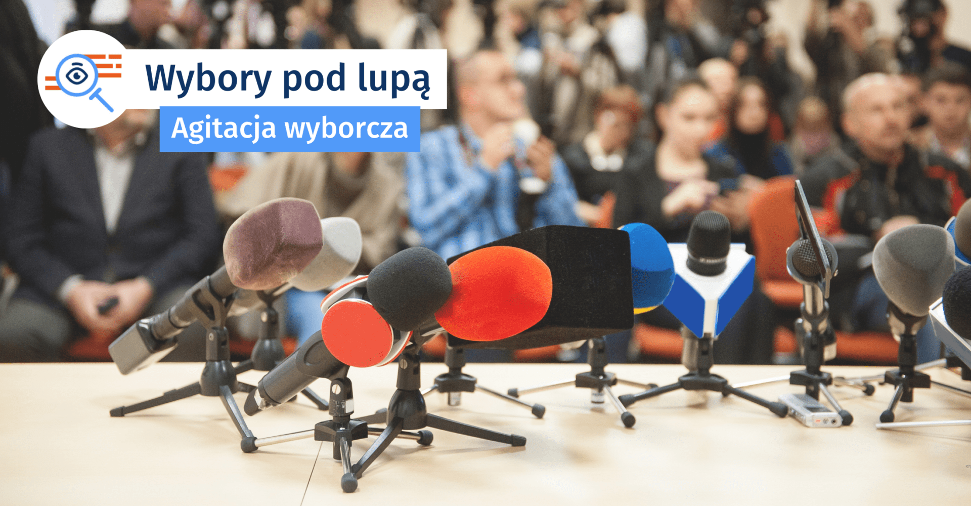 Sala konferencyjna. Na pierwszym planie stojące na stole mikrofony. W tle nieostra zgromadzona publiczność.