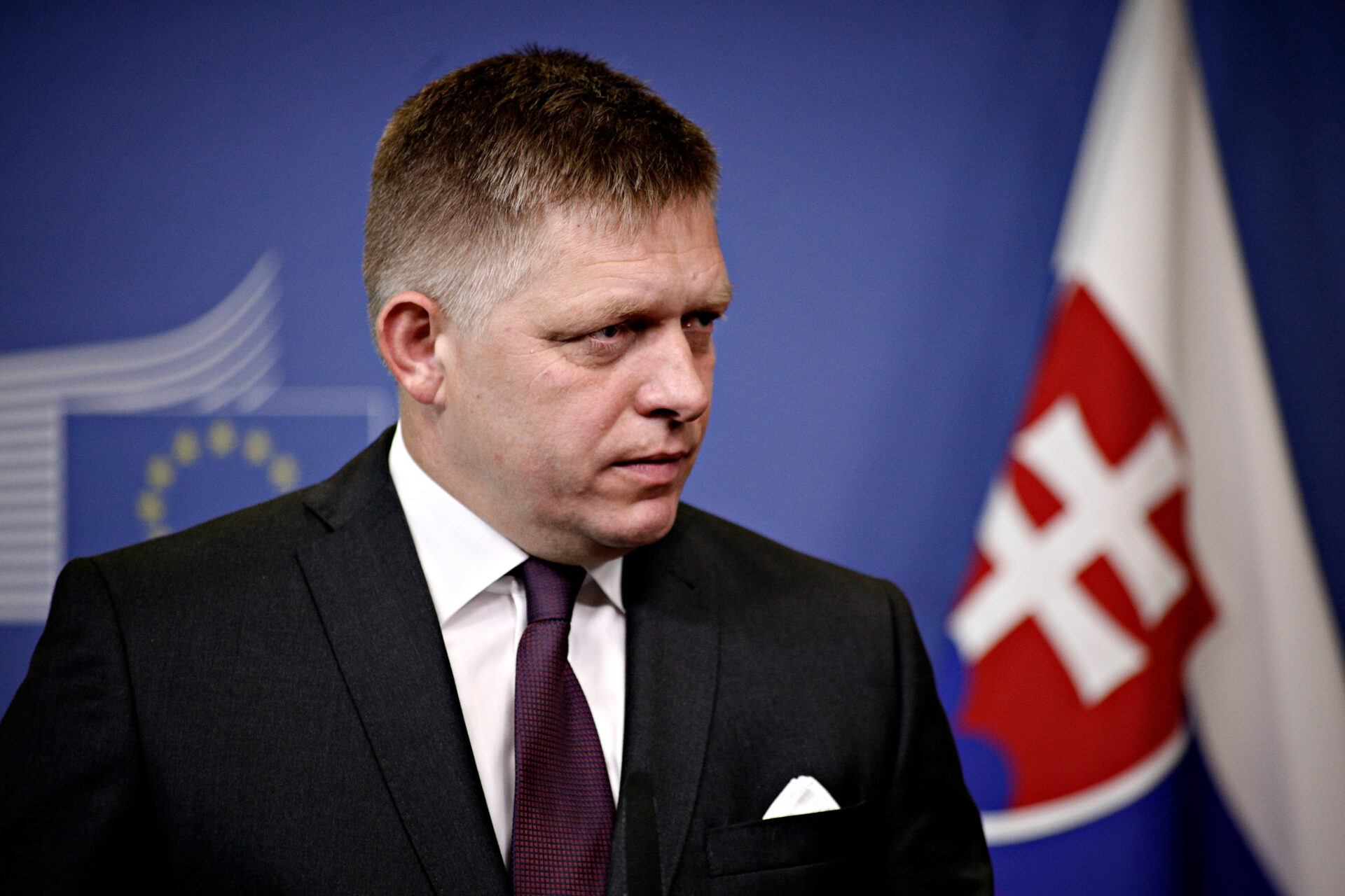 Robert Fico w czarnym garniturze i bordowym krawacie, patrzy w prawo. Na tle logotyp Komisji Europejskiej, po prawej stronie flaga Słowacji.