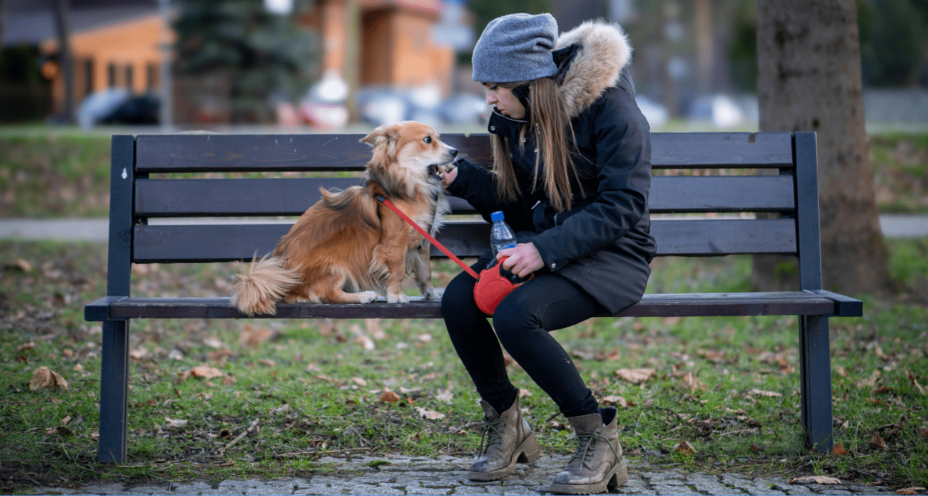Kobieta ubrana w zimową kurtkę z szarą czapką na głowie siedzi na ławce ze swoim psem. W ręce trzyma ona czerwoną smycz oraz butelkę wody.