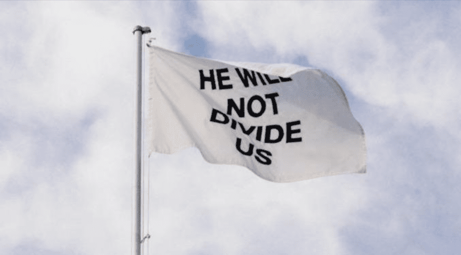 Zdjęcie białej flagi z napisem „We will not divide us”, która powiewa na wietrze. W tle widać zachmurzone niebo.