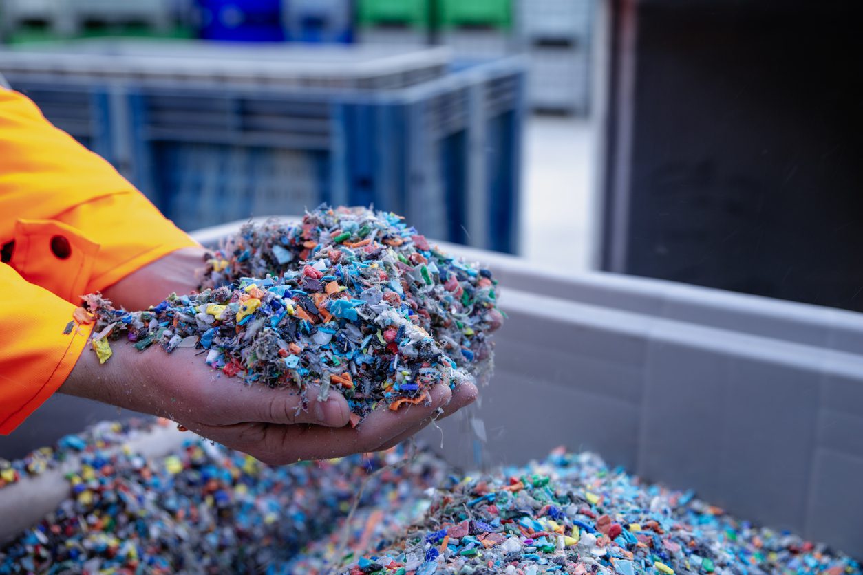 Dłonie pełne pokruszonego plastiku nad kontenerem z mikroplastikiem. W tle inne szare kontenery.