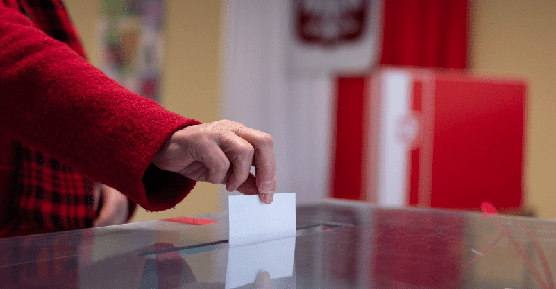 na grafice widzimy dłoń wrzucającą kartę wyborczą do urny