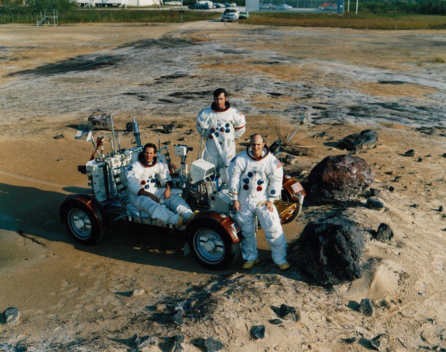 Kolorowe zdjęcie przedstawiające trzech astronautów misji Apollo 16 w trakcie ćwiczeń, stojących obok łazika kosmicznego ze zdjętymi hełmami. W górze kadru widać samochody i fragment parkingu.