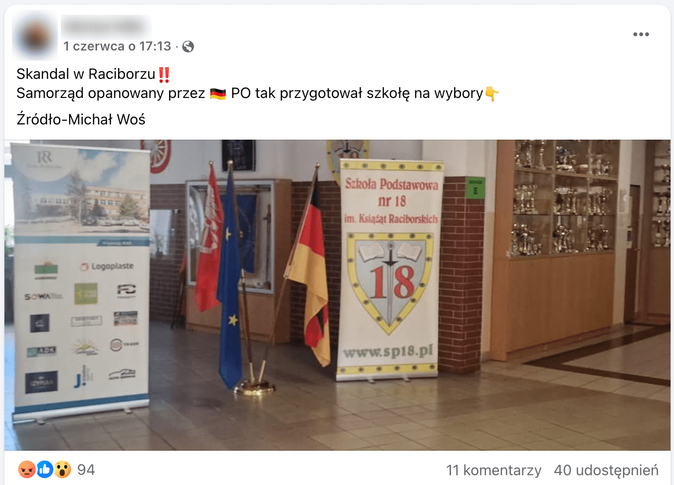 Zrzut ekranu posta na Facebooku. Załączone zdjęcie przedstawia korytarz szkolny. Pod ścianą stoi gablota z pucharami. Na środku ustawiono dwa banery oraz trzy flagi: Unii Europejskiej, Niemiec. Trzecia flaga ma czerwoną barwę oraz biały emblemat, trudno określić, co przedstawia. 
